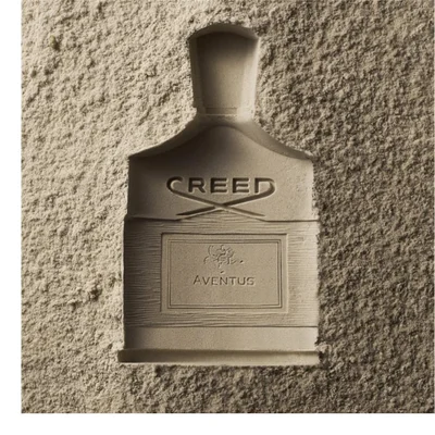 A dark bottle of Creed Absolu Aventus perfume with a rider on a horse logo, marked 75 ML 2.5FL.OZ., is tilted over two citrus fruits.