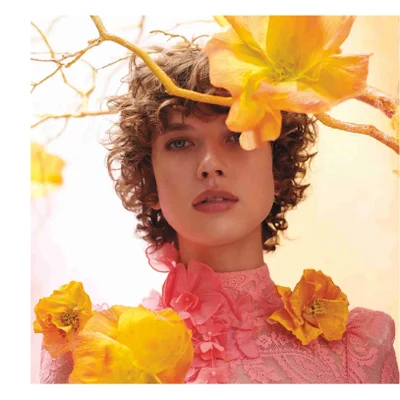 A young woman with curly brown hair looks directly forward, adorned with large yellow paper-like flowers and golden branches around her head and shoulders, wearing a pink lace dress with floral embellishments.