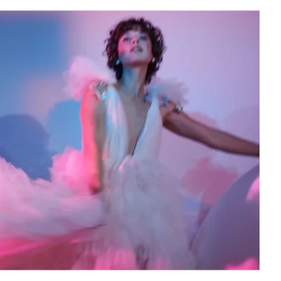 A person with dark curly hair and a white, deep V-neck ruffled dress with sparkling shoulder adornments looks up, captured with motion blur against a background of blue, pink, and purple light.