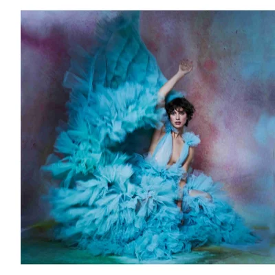 A woman in a voluminous teal ruffled gown poses with her arm raised against a colorful mottled background.