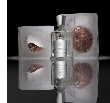 A Creed perfume bottle is shown standing between two ice blocks with pieces of coconut frozen inside, reflected on a dark surface below, with the label reading CREED VIRGIN ISLAND WATER DE PÈRE EN FILS DEPUIS 1760 FROM FATHER TO SON SINCE 1760 100ML 3.33 FL.OZ.