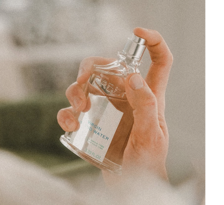 A hand holds a glass bottle of Creed Virgin Island Water perfume showing the label with text VIRGIN ISLAND WATER, FRANCAIS DEPUIS 1760 ENGLISH SINCE 1760, and 3.33 FL. OZ.