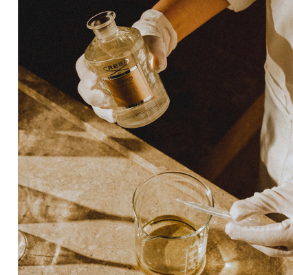 A person in white gloves pours liquid from a glass bottle labeled CREED ESIME IMPERIAL into a beaker.