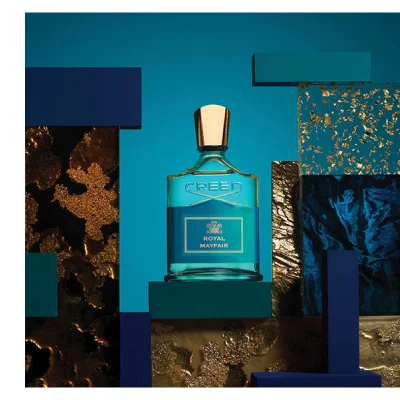 A Creed Royal Mayfair perfume bottle is displayed on a teal block surrounded by abstract blocks in blue, teal, gold texture, and dark blue texture against a blue background, with text reading CREED ROYAL MAYFAIR.