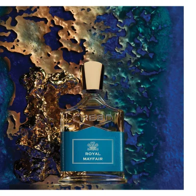 A perfume bottle with a blue label stands next to a gold object against a textured blue and gold background with text reading CREED ROYAL MAYFAIR.