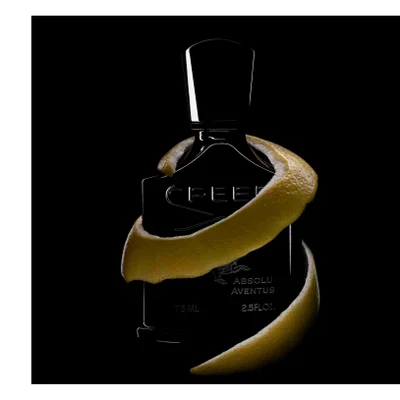 A dark perfume bottle with a citrus peel wrapped around it against a black background, showing the text CREED ABSOLU AVENTUS 75 ML 2.5 FL.OZ.