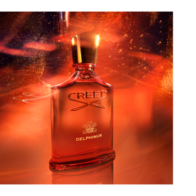 A red perfume bottle with a gold cap is shown against a fiery orange background with gold sparkles, with the text CREED DELPHINUS visible on the bottle.