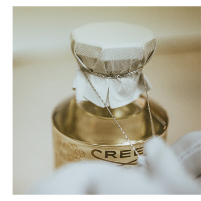 Close-up of a golden bottle, likely perfume, with a cloth-covered cap tied with a string, showing part of the bottles label with text CREED 1760.