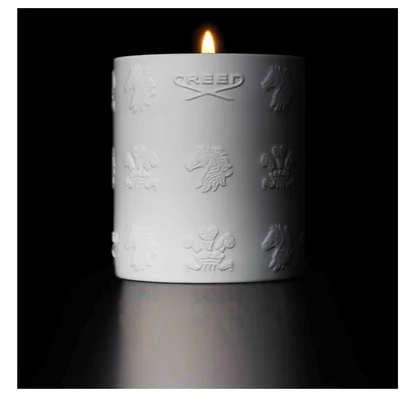 A lit white candle with embossed designs and the text CREED sits on a reflective surface in a dark setting