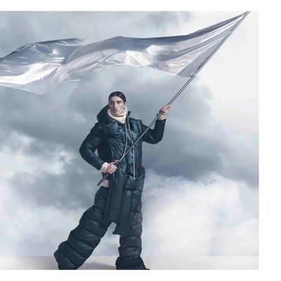 A person wearing a dark green puffer jacket and pants with a cream scarf holds a pole with a large metallic flag against a cloudy sky.