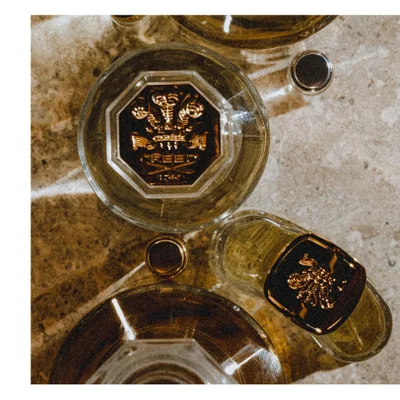 Close-up of a golden bottle, likely perfume, with a cloth-covered cap tied with a string, showing part of the bottles label with text CREED 1760.