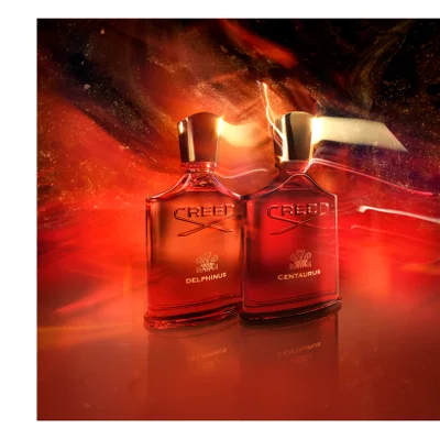 Two Creed perfume bottles, a red one with text CREED CENTAURUS and an orange one with text CREED DELPHINUS, are reflected on a surface against a swirling red and orange background.