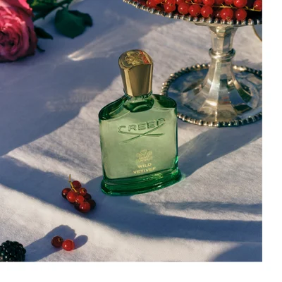 A green perfume bottle with a gold cap rests on a white plate surrounded by red currants and green grass, displaying the text CREED and WILD VETIVER.