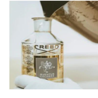 A white-gloved hand holds a Creed Aventus Cologne bottle as liquid is poured into it from a dark container above, with the text CREED and AVENTUS COLOGNE visible on the bottles label.