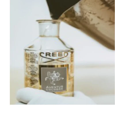 A hand in a white glove holds a clear glass cologne bottle with gold liquid, into which a dark container is pouring more liquid, and the bottle label displays CREED and AVENTUS COLOGNE.