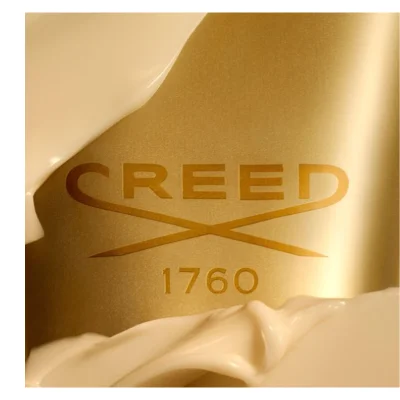 A close-up of a gold surface partially covered by a swirling light cream substance, featuring the embossed text CREED and 1760.