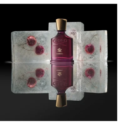 A dark magenta perfume bottle with a gold cap stands centered, flanked by two ice blocks with embedded red berries, all reflected on a glossy surface, and the bottle displays the text CREED and CARMINA.
