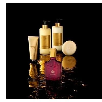 Five luxury Creed Carmina beauty products are arranged on a dark, rippling surface with golden reflections, featuring a dark red perfume bottle, a gold tube of hand cream, two cream-colored pump bottles for hand and body wash and lotion, and a round soap bar; visible text on the products includes CREED 1760, CARMINA, HAND CREAM, CRÈME MAINS, HAND & BODY WASH, LAVANT MAINS ET CORPS, HAND & BODY LOTION, and LAIT MAINS ET CORPS.