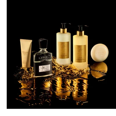 A striking display of five Creed Aventus fragrance and body products, including a black perfume bottle, a golden hand cream tube, two tall golden pump bottles, and a creamy bar of soap, emerges from golden liquid splashes on a reflective black background, with visible text including CREED, 1760, AVENTUS, HAND CREAM, CREME MAINS, HAND & BODY LOTION, LOTION MAINS ET CORPS, HAND & BODY WASH, and LAVANT LIQUIDE MAINS ET CORPS.