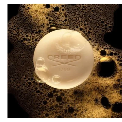 A round, light-colored bar of Creed soap with the text CREED and 1760 embossed on it, surrounded by golden-brown foam and bubbles on a dark background.