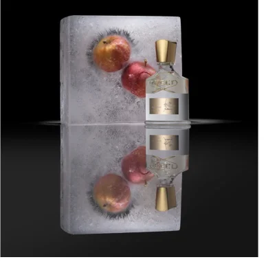 A clear perfume bottle with a gold cap and label is positioned next to a rectangular block of ice that has three red and yellow apples frozen inside it, with both items and their reflections visible on a dark surface, and the bottles label reads CREED AVENTUS.