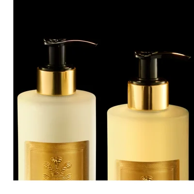 A collection of five gold and cream colored Creed Aventus For Her beauty products, including a perfume bottle, two pump bottles, a hand cream tube, and a bar of soap, are arranged on a reflective black surface with golden liquid ripples, bearing the visible text Creed 1760, Aventus For Her, Hand Cream, Crème Mains, Hand & Body Wash, Savon Liquide Mains et Corps, Hand & Body Lotion, Lotion Mains et Corps.