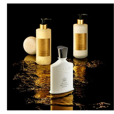 A black Creed perfume bottle, a cream-colored lotion bottle with a gold pump, and a round cream-colored bar of soap from the Green Irish Tweed line are displayed on a reflective black surface with shimmering gold liquid; the perfume bottle reads CREED, GREEN IRISH, TWEED, the lotion bottle reads CREED, 1760, GREEN IRISH TWEED, HAND & BODY WASH, SAVON LIQUIDE MAINS ET CORPS, and the soap reads CREED, 1760.