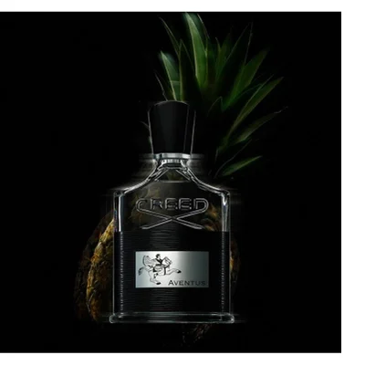 A clear glass bottle of Creed Aventus cologne with a black ribbed band and a silver label depicting a horse and rider is displayed in front of a pineapple on a dark background, showing text CREED AVENTUS.