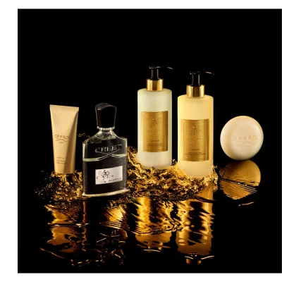 Five Creed Aventus luxury bath and body products, including a gold hand cream tube, a black-capped perfume bottle, a white pump lotion bottle, a yellow pump body wash bottle, and a round bar of soap, are displayed amidst golden liquid splashes on a reflective black surface. Visible text: CREED 1760 AVENTUS HAND CREAM CREME MAINS CREED AVENTUS CREED 1760 AVENTUS HAND & BODY LOTION LOTION MAINS ET CORPS CREED 1760 AVENTUS HAND & BODY WASH LAVANT LIQUIDE MAINS ET CORPS CREED 1760.