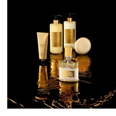Five gold-accented Creed Aventus for Her beauty products are artfully arranged on a dark, reflective surface with golden liquid ripples, showcasing visible text that reads CREED 1760 AVENTUS FOR HER HAND CREAM CRÈME MAINS on the hand cream tube, CREED 1760 AVENTUS FOR HER SAVON LIQUIDE BODY & HAND WASH on the liquid soap bottle, CREED 1760 AVENTUS FOR HER HAND & BODY LOTION LAIT ONCTUEUX MAINS ET CORPS on the body lotion bottle, CREED 1760 on the bar of soap, and CREED AVENTUS FOR HER with a warrior on horseback graphic on the perfume bottle.
