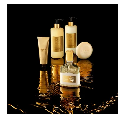 A collection of five gold and cream colored Creed Aventus For Her beauty products, including a perfume bottle, two pump bottles, a hand cream tube, and a bar of soap, are arranged on a reflective black surface with golden liquid ripples, bearing the visible text Creed 1760, Aventus For Her, Hand Cream, Crème Mains, Hand & Body Wash, Savon Liquide Mains et Corps, Hand & Body Lotion, Lotion Mains et Corps.