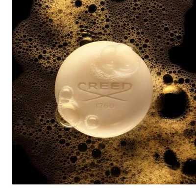 A round, creamy white soap bar with bubbles on its surface rests in golden-brown foam, displaying the embossed text CREED 1760. Image 1 - A round, creamy white soap bar with clear lather bubbles rests on a dark golden-brown bubbly foam, featuring the embossed text CREED 1760.