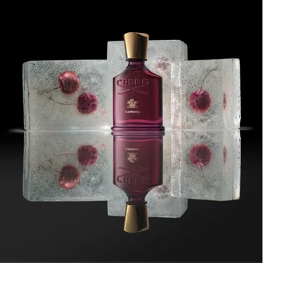 A dark reddish-purple Creed Carmina perfume bottle with a gold cap stands centered between ice blocks containing frozen red berries, reflecting on a dark surface. Image 2 - A dark reddish-purple perfume bottle with a gold cap is centered among ice blocks containing frozen red cherries, reflected on a dark surface, displaying the text CREED and CARMINA.