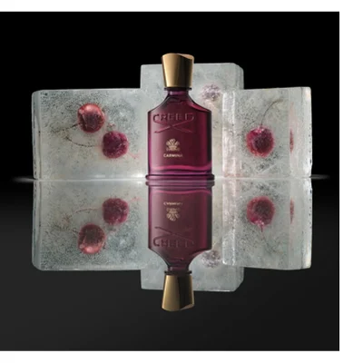 A deep red perfume bottle with a gold cap stands on a reflective surface against a dark background, flanked by four ice blocks containing red frozen cherries, with visible text CREED and CARMINA on the bottle.